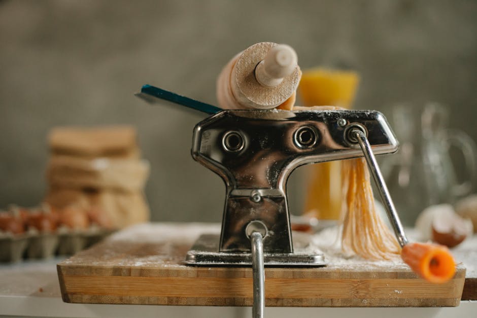 Stainless modern pasta maker instrument with handle and cut dough placed on table with products on blurred background in kitchen
